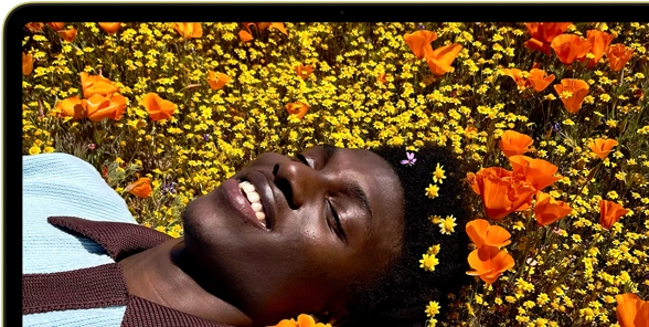 MacBook Neo, citrus color, screen shows a smiling person lying in a sunny field of flowers