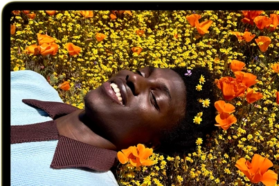 MacBook Neo, citrus color, screen shows a smiling person lying in a sunny field of flowers