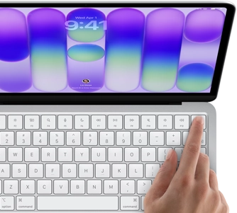 MacBook Neo, silver color, showing login screen, person's finger on Touch ID sensor in top-right corner of keyboard