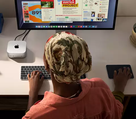 A person typing at a desk with a Mac mini, Studio Display connected, Magic Keyboard, Magic Trackpad, various items