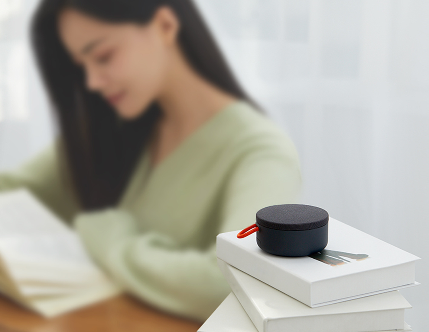 Mi Portable Bluetooth Speaker свири върху купчина книги, докато жена чете до прозореца
